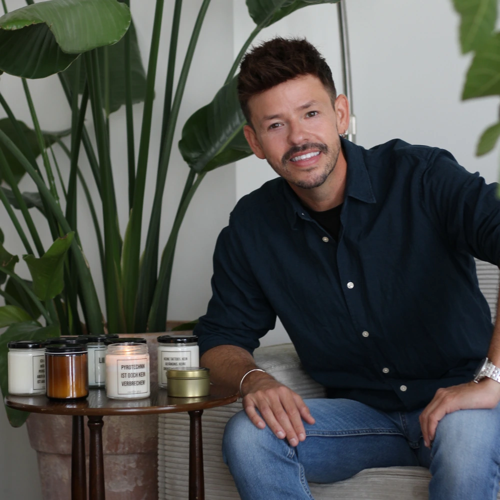 Michael Murphy, founder of Candle Guy, sitting next to a table with handmade scented candles featuring cheeky quotes in glass jars.